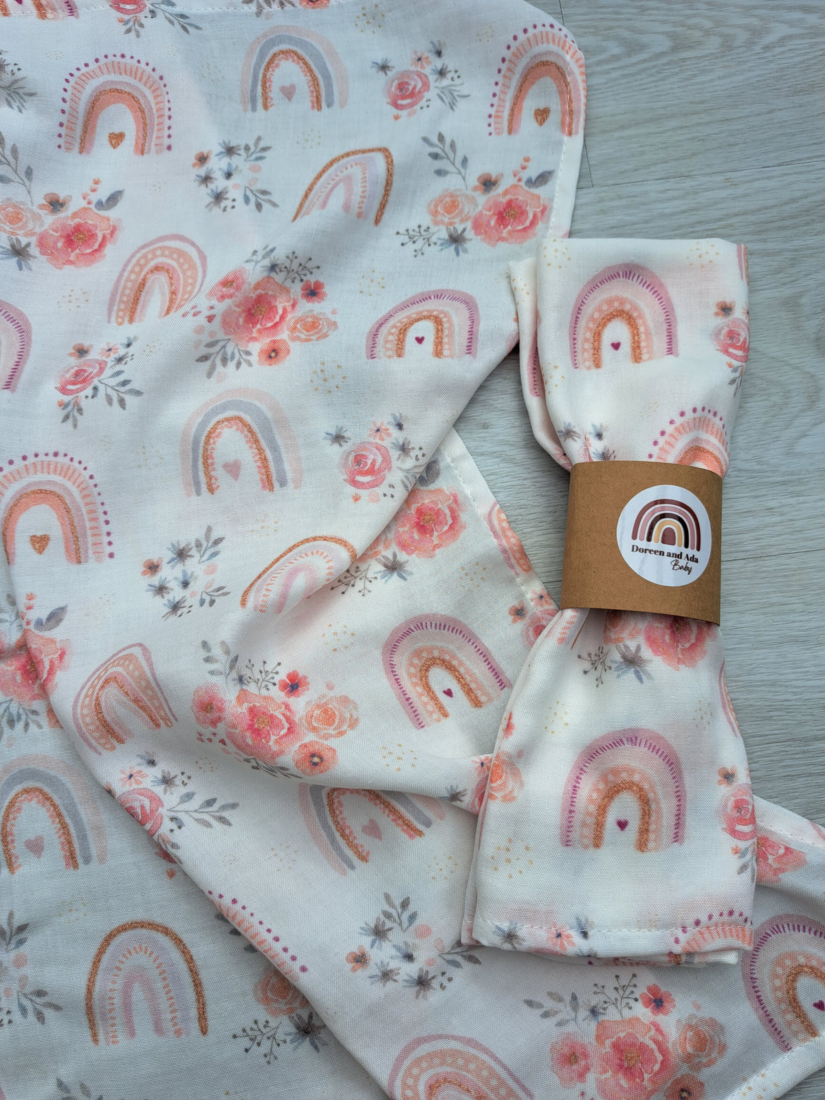 Rainbows and roses muslin square rolled with Doreen and Ada Baby band on unfolded muslin on wooden floor showing watercolour rainbow and pink floral repeat