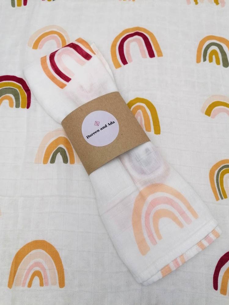 Boho rainbow muslin square rolled with Doreen and Ada band on matching rainbow print background showing warm earthy boho arched rainbows in mustard yellow sage green crimson and blush pink on white cotton from above