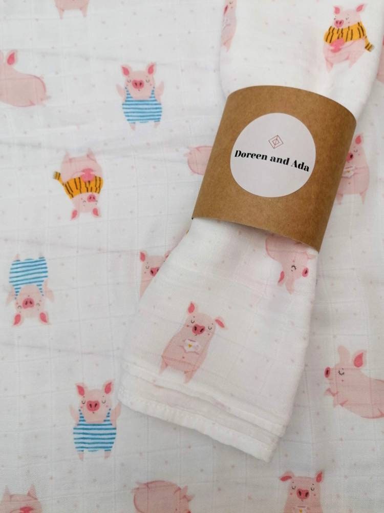 Pig muslin square rolled with Doreen and Ada band on matching pig print background showing close-up of watercolour piglets in blue striped dungarees and yellow and orange striped tops on white cotton