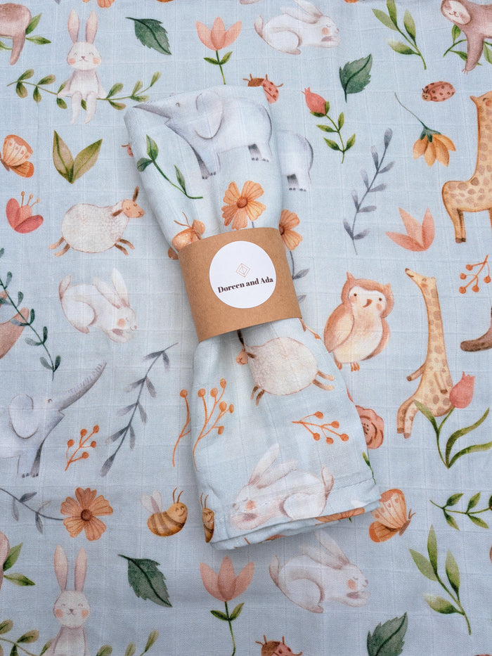 A light blue cotton and bamboo muslin square with an animal pattern, displayed with a brand tag showing the product name and price.