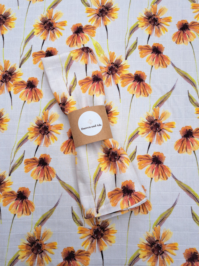 A muslin square with an orange floral pattern on a light background, displayed with a brand tag on the side.