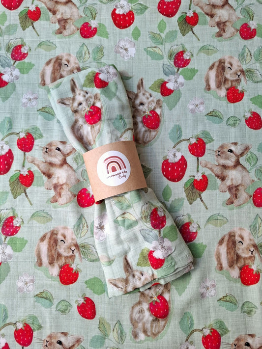 Muslin square with a mint background featuring bunnies holding strawberries surrounded by leaves and flowers, folded with an eco packaging label