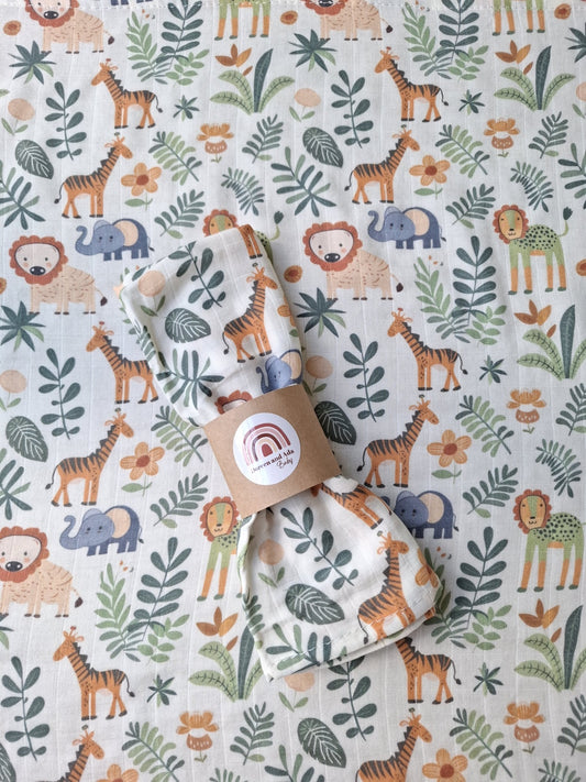 Muslin square featuring a printed pattern of giraffes, lions and elephants with green leaves and orange flowers on soft cotton fabric