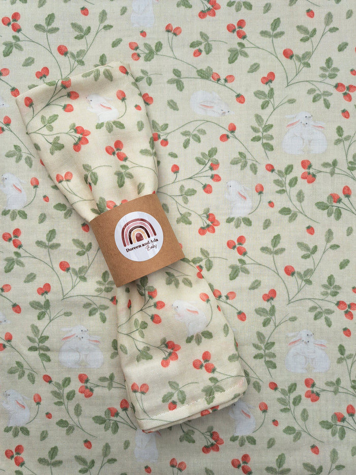 A folded beige muslin blanket with a floral pattern and rabbit motifs, presented with a brand tag.