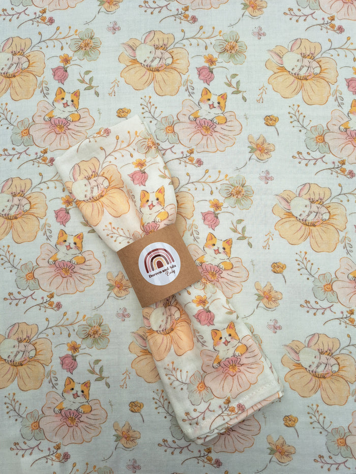 A floral muslin square with a pattern of cats and rabbits, presented with a brown paper label featuring a white circular logo.