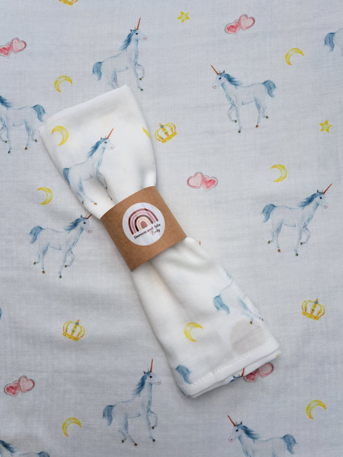 A folded white muslin blanket with a unicorn and moon pattern, presented with a brand tag showing a unicorn and the text 'organic cotton'.