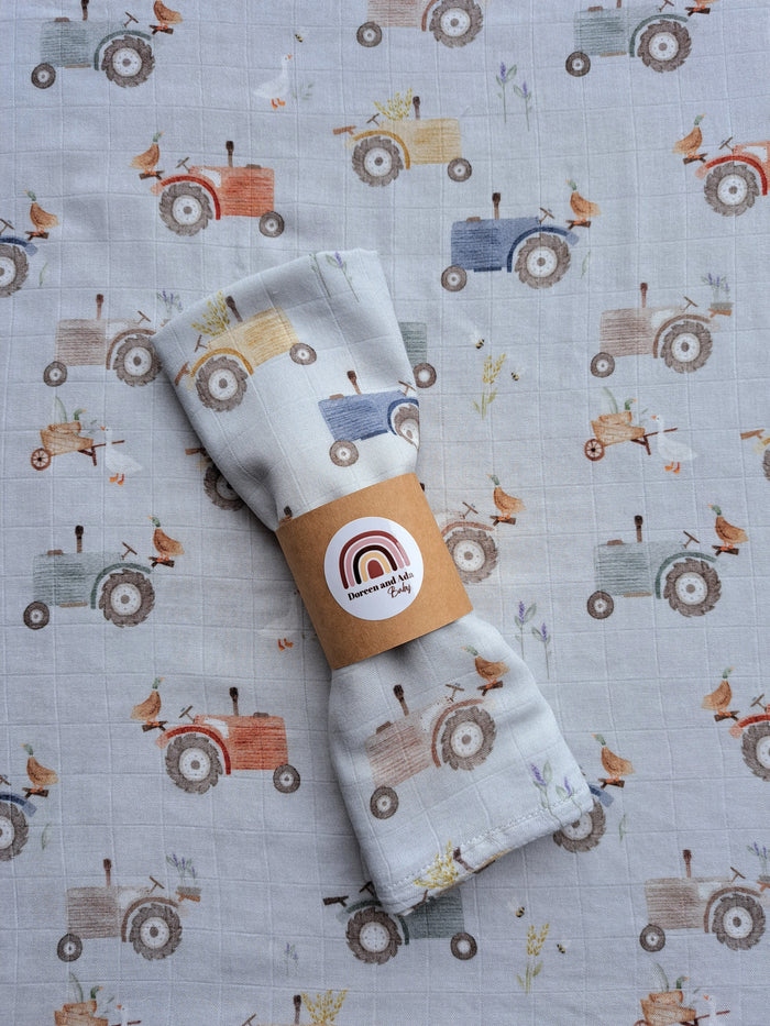 A swaddling blanket with a tractor and farm animals pattern, displayed on a patterned background.