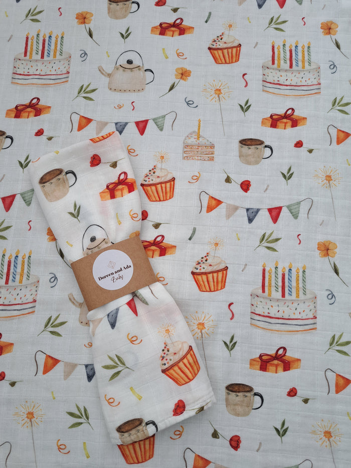 A muslin square with a printed pattern featuring various tea-related items like cups, cakes, and decorations, with a bear-like character drinking tea from a cup.