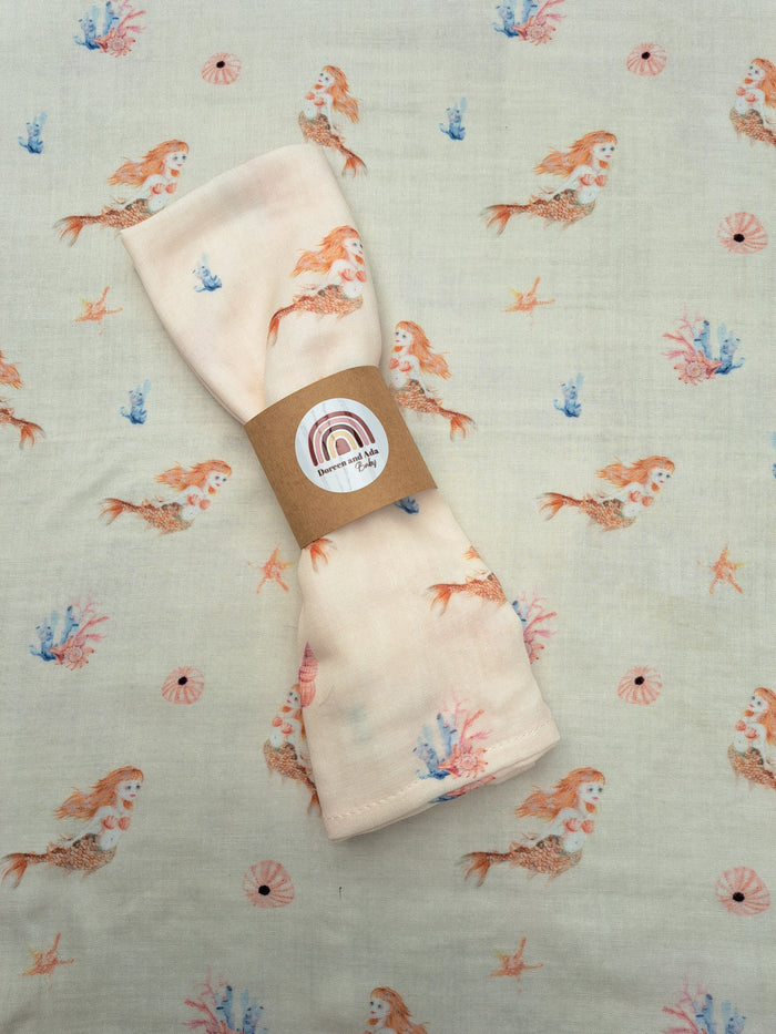 A folded beige muslin square blanket with a printed mermaid tail and fin design, placed on a similar patterned background.