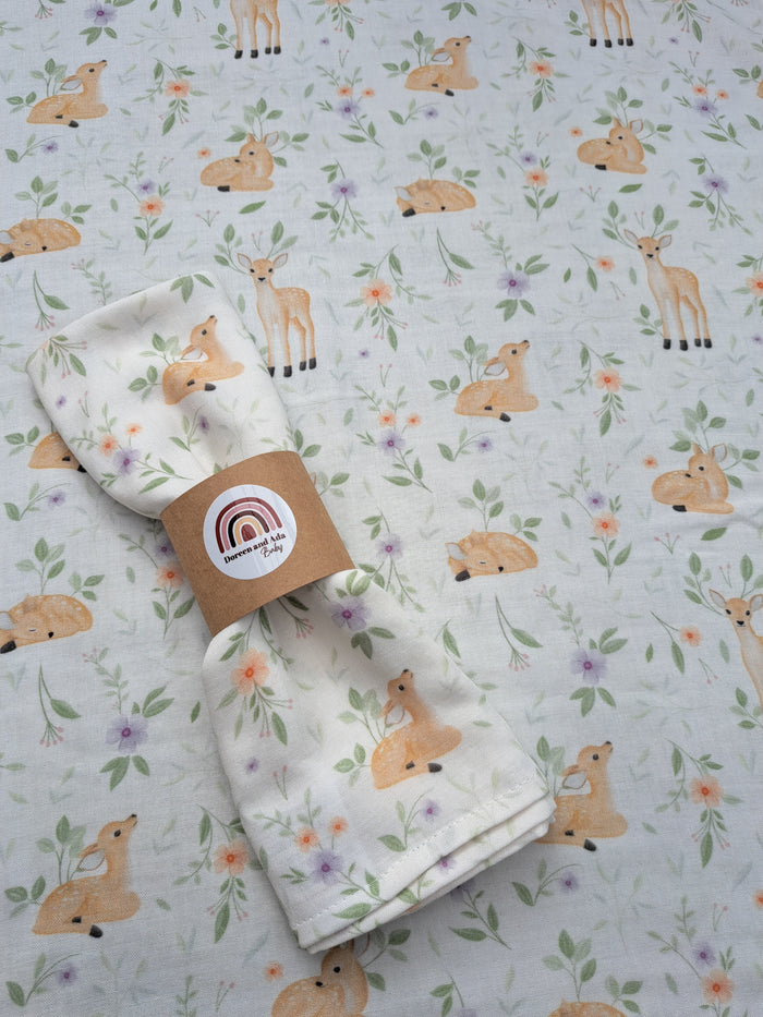 A folded beige muslin blanket with a floral pattern and a deer motif, presented with a brand tag.