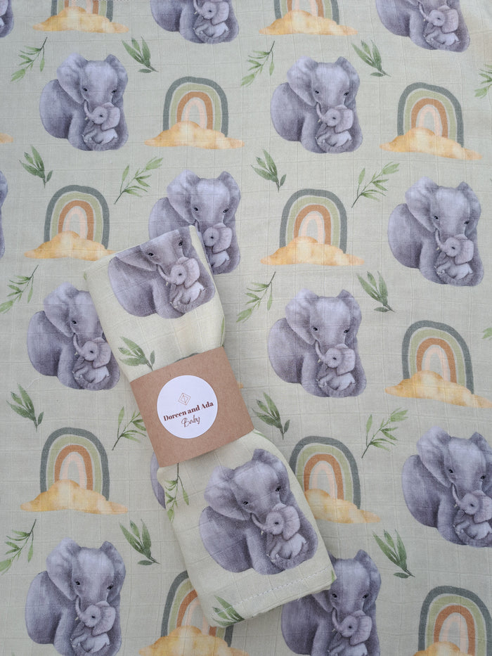 A swaddling blanket with a pattern of gray elephants in various poses and colors on a beige background, with a rainbow motif in the background.