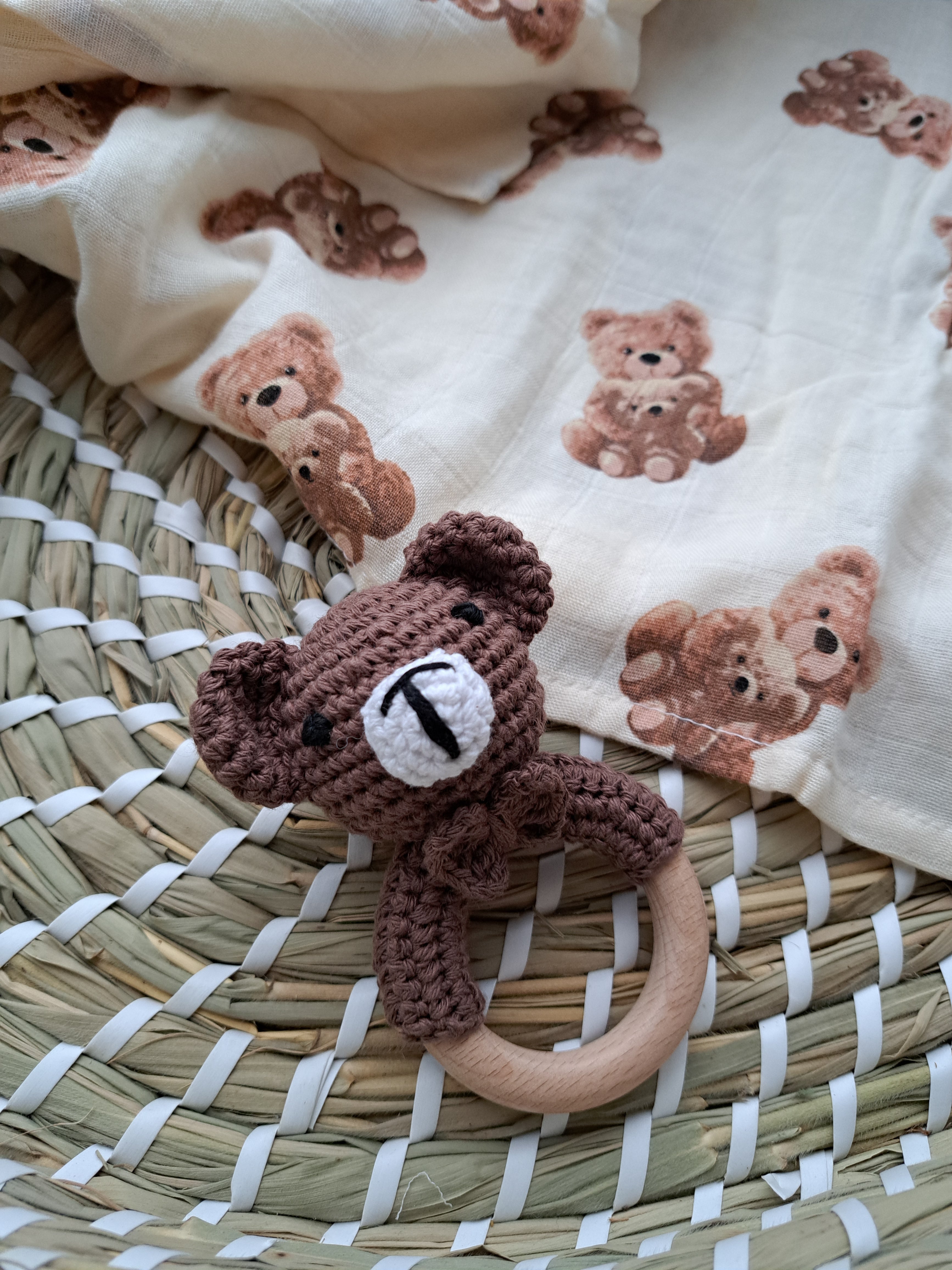 A brown bear-shaped teether rattle with a bell, made from smooth beech wood and soft cotton, placed next to a beige fabric with bear prints.