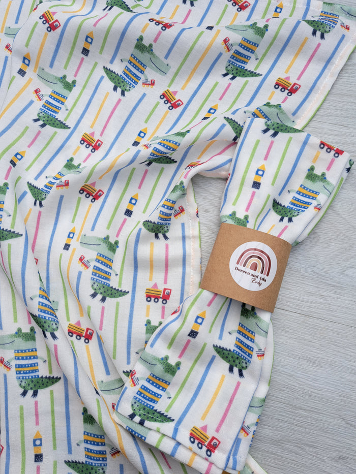 A muslin square baby blanket with a striped pattern featuring colorful crocodile and building block motifs, displayed with its packaging.