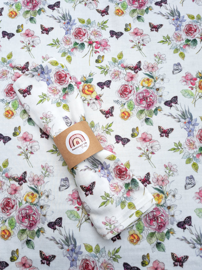 A folded white muslin square blanket with a floral and butterfly pattern, presented on a background with a similar pattern. It is packaged in a brown paper with a logo and a small tag showing a butterfly.