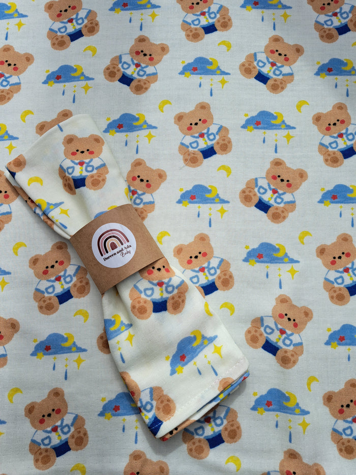 A folded beige muslin blanket with a colorful teddy bear pattern, presented with a brand tag attached.