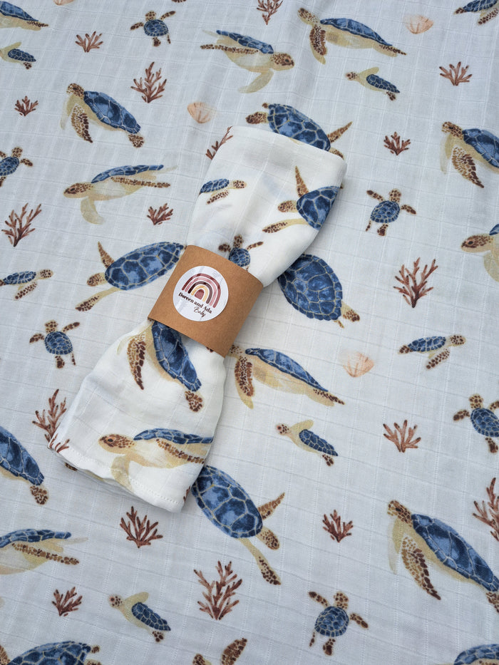 A muslin square with a pattern of turtles and sea waves on a light background, displayed on a flat surface.
