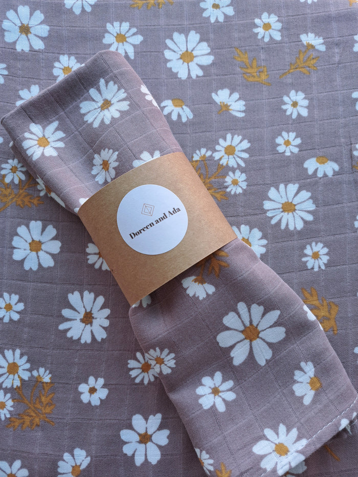 A lavender muslin square with daisy floral print, wrapped with a band and a tag showing the brand name.